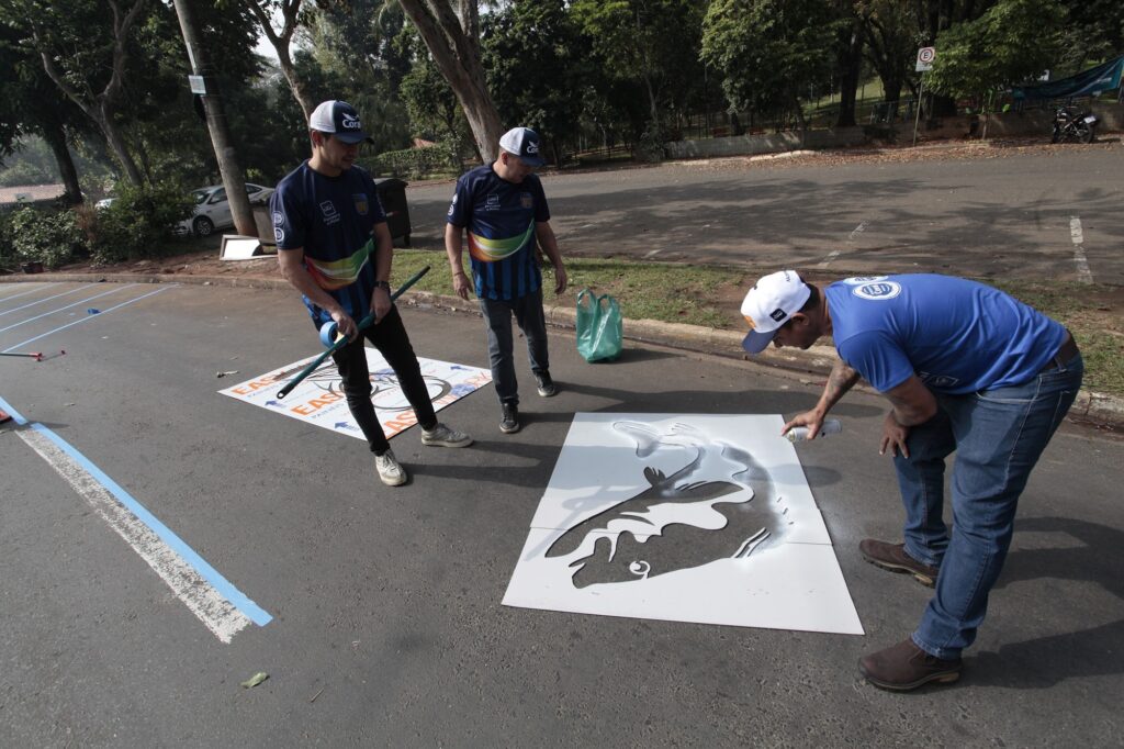A imagem mostra três homens em um ambiente externo, possivelmente um estacionamento ou uma área pavimentada em um parque, realizando uma atividade de pintura no chão. Dois homens estão de pé, um deles segurando uma fita adesiva azul em um bastão longo, enquanto o outro observa. O terceiro homem está agachado, aplicando tinta spray através de um estêncil em formato de peixe no asfalto. Todos os homens vestem camisetas azuis com o logo da marca "Coral" e bonés brancos, indicando que fazem parte de uma equipe ou evento organizado pela marca. No chão, há fitas azuis coladas, provavelmente para demarcar áreas ou auxiliar na aplicação da pintura. Também é possível ver algumas árvores ao fundo, sugerindo que o local é ao ar livre e em um ambiente com vegetação. A cena transmite uma sensação de trabalho colaborativo e cuidado na execução da pintura, que parece ser parte de uma ação de sinalização ou decoração do espaço público. A imagem é clara e bem iluminada, destacando o momento da pintura em processo.