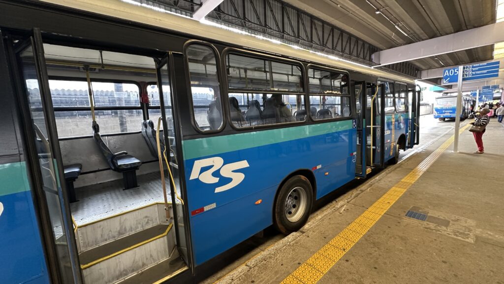 A imagem mostra uma estação de ônibus urbana durante o dia. Em primeiro plano, vemos a lateral de um ônibus azul e verde da empresa "RS" parado com as portas abertas, revelando o interior com assentos escuros e piso metálico. * À direita, uma passageira caminha pela plataforma, que possui faixas de sinalização tátil amarela no chão. * Ao fundo, é possível observar outro ônibus estacionado e placas de sinalização suspensas indicando números de linhas, como "A05", "120", "406" e "430", sugerindo um terminal de transporte público movimentado. O ambiente é coberto por uma estrutura metálica de teto alto.
