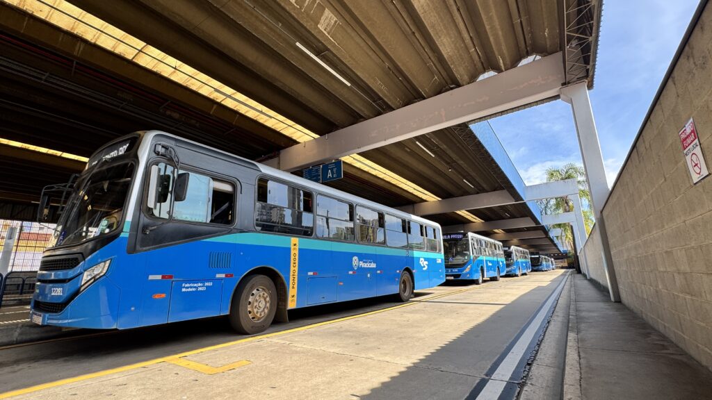 A imagem mostra um terminal de ônibus urbano coberto, com estrutura ampla e moderna, onde vários ônibus azuis estão organizados em fila nas plataformas. Em primeiro plano, há um ônibus destacado, estacionado ao lado da plataforma sinalizada, com portas e janelas amplas e identificação do sistema de transporte municipal. Ao fundo, outros ônibus seguem alinhados, reforçando a rotina operacional do terminal. O espaço é coberto por uma grande estrutura metálica, com aberturas que permitem a entrada de luz natural e criam contraste entre áreas de sombra e de sol. À direita, vê-se um corredor de circulação para pedestres ao longo de um muro de concreto, além de sinalizações de orientação e regras do local.