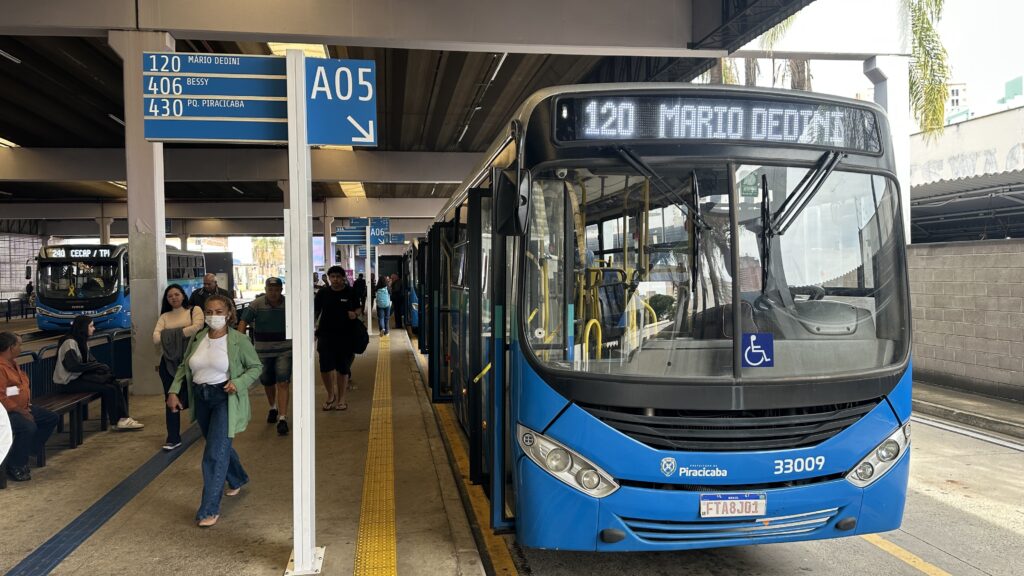 A imagem mostra uma cena em um terminal de ônibus urbano na cidade de Piracicaba, Brasil. Em destaque, há um ônibus azul moderno parado na plataforma A05, com o letreiro eletrônico indicando a linha "120 MARIO DEDINI". O ônibus possui o símbolo de acessibilidade para cadeirantes na parte frontal e a placa FTA8J01. Ao redor, algumas pessoas caminham pela plataforma, incluindo uma mulher na frente usando máscara facial branca, jaqueta verde e jeans. O terminal é coberto, com pilares de concreto e piso com faixa tátil amarela para acessibilidade. Acima da plataforma, há placas azuis indicando as linhas de ônibus disponíveis, como 120, 406 e 430. Ao fundo, é possível ver outro ônibus e mais estruturas do terminal. A iluminação é natural, sugerindo que a foto foi tirada durante o dia. O ambiente transmite a rotina do transporte público na cidade, mostrando a infraestrutura e a movimentação típica de passageiros.