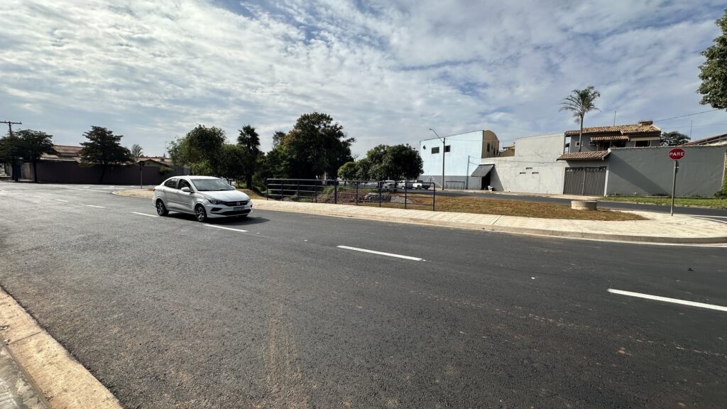 A imagem mostra uma cena urbana durante o dia, com céu parcialmente nublado e iluminação natural. No centro da imagem, um carro branco está trafegando por uma rua asfaltada recentemente, com faixas brancas pintadas no asfalto. À direita da rua, há um meio-fio seguido por uma pequena área gramada e um muro baixo que delimita uma área residencial, onde se vê uma casa de cor azul clara e outra estrutura de concreto. Também à direita, próximo ao meio-fio, está uma placa de trânsito vermelha com a palavra "PARE", indicando que os veículos devem parar naquele ponto. Ao fundo, há árvores e outros elementos típicos de uma área urbana, além de alguns outros carros à distância. A imagem transmite uma sensação de tranquilidade e organização no trânsito local.
