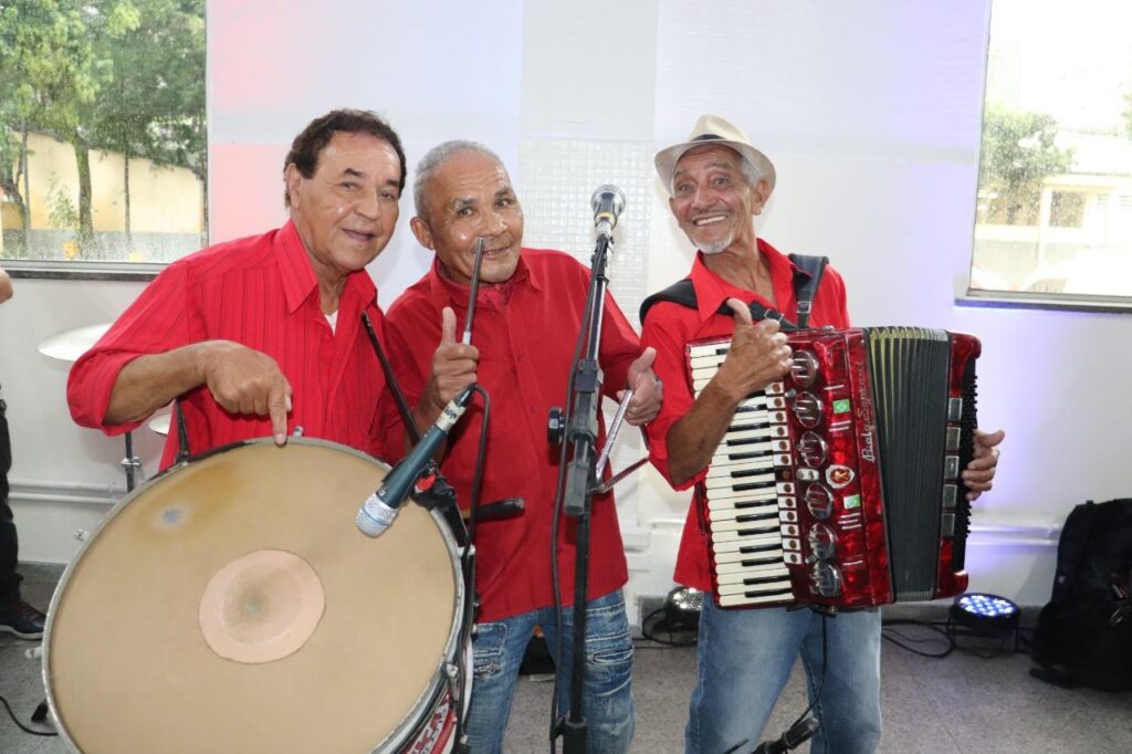 Esta foto mostra um trio musical de senhores posando alegremente para a câmera. Todos estão vestindo camisas sociais vermelhas de manga curta e jeans. À esquerda: Um homem segura um zabumba (um instrumento de percussão típico do forró). Ao centro: Um homem segura uma baqueta e faz um sinal de "joinha" com a mão. À direita: Um homem usa um chapéu, toca uma sanfona vermelha e também faz um sinal de "joinha". Eles parecem estar em um ambiente interno, possivelmente se preparando para ou encerrando uma apresentação musical, já que há microfones e equipamentos de som visíveis ao redor deles.