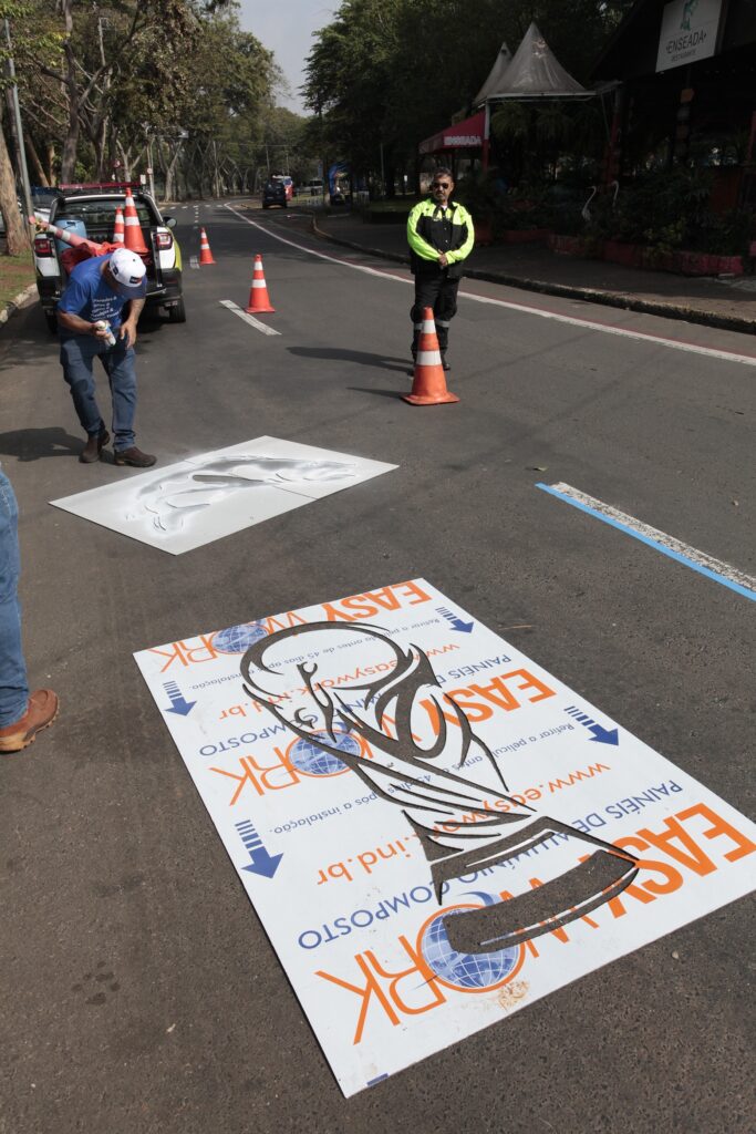 A imagem mostra uma cena em uma rua pavimentada durante o dia, onde duas pessoas estão pintando estênceis no asfalto. No primeiro plano, há um grande estêncil retangular com o logotipo da empresa "EASY WORK" e a imagem da taça da Copa do Mundo da FIFA. Um homem, vestido com uma camiseta azul e boné branco, está usando um spray para aplicar tinta através de um estêncil em forma de peixe. Outro homem, usando uma jaqueta de alta visibilidade amarela e preta, observa o processo. A rua está parcialmente bloqueada com cones de trânsito, e ao fundo, há um veículo branco estacionado. Também é possível ver árvores e um prédio com a placa "ENSEADA RESTAURANTE". A iluminação é natural, com sombras definidas no chão, indicando um dia claro e ensolarado. A cena parece ser parte de um trabalho de sinalização ou decoração urbana.