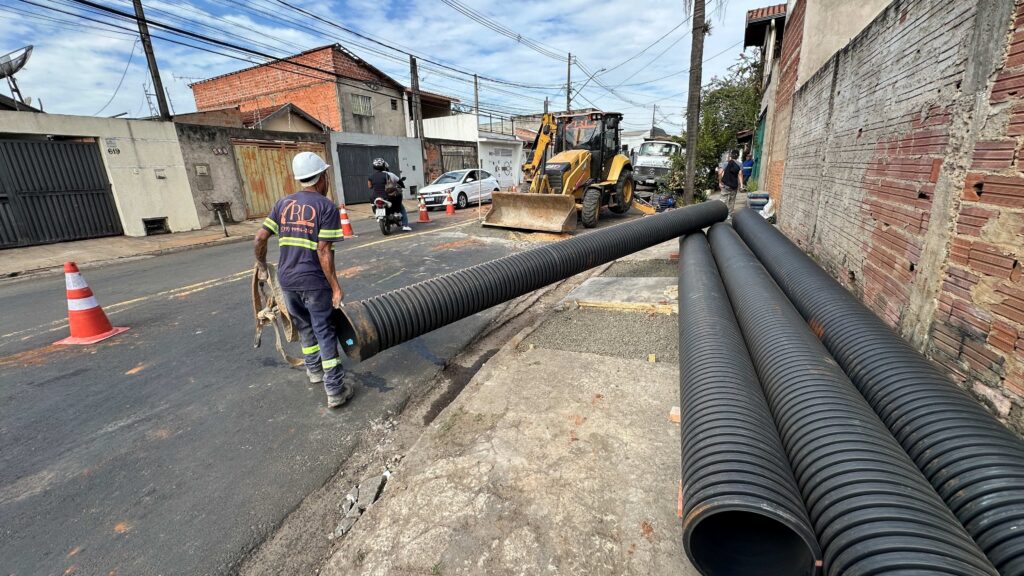 A imagem mostra uma cena de rua durante o dia, em uma área residencial. Na frente, um trabalhador da construção civil, usando capacete branco e uniforme azul escuro com as letras "RD", está segurando uma tubulação preta corrugada, provavelmente para instalação subterrânea. No chão, ao lado, há outras tubulações semelhantes. No meio da rua, há uma máquina amarela de construção, uma retroescavadeira da marca CAT, que parece estar parada. Também é possível ver uma motocicleta passando pela rua. As casas ao redor são de alvenaria, com muros e portões de metal. O ambiente está iluminado pela luz do dia, com céu parcialmente nublado. A cena sugere que está em andamento um serviço de infraestrutura, como instalação ou manutenção de rede de esgoto, drenagem ou cabos subterrâneos.