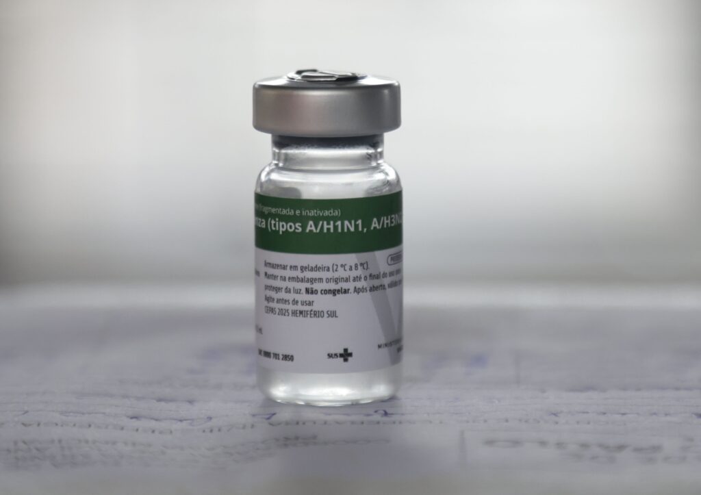 A imagem mostra um frasco de vacina contra a influenza, em posição vertical e com foco nítido. O frasco é de vidro transparente, com tampa metálica prateada. Ele contém um líquido claro e possui um rótulo branco com detalhes em verde. No rótulo, é possível ler informações em português, indicando que a vacina é contra os tipos A/H1N1 e A/H3N2 do vírus da gripe, destinada para a temporada de 2025 do hemisfério sul. Também há instruções de armazenamento, recomendando manter a vacina refrigerada entre 2 °C e 8 °C, protegida da luz e na embalagem original até o uso, além de não congelar o produto. A imagem transmite um ambiente clínico e profissional, com fundo desfocado em branco, sugerindo um contexto de saúde pública.