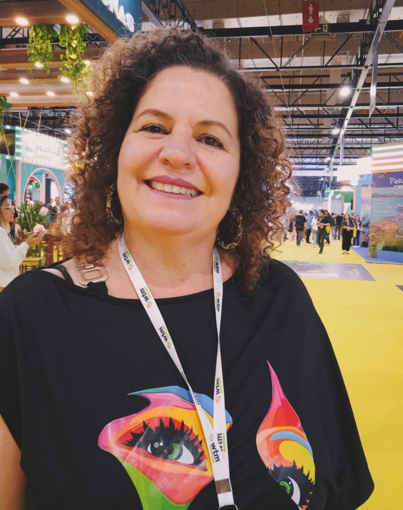 A imagem mostra uma mulher sorridente em um ambiente que parece ser uma feira ou conferência, possivelmente relacionada a tecnologia ou negócios, dado o crachá com a sigla "wtm" que ela usa no pescoço. Ela tem cabelo castanho cacheado na altura dos ombros e veste uma blusa preta com uma estampa colorida que lembra um olho estilizado com padrões em tons de arco-íris. Ela também usa brincos grandes e coloridos. O fundo é um espaço amplo com piso amarelo e várias pessoas e estandes, sugerindo um evento movimentado. A iluminação é clara e destaca bem a pessoa em primeiro plano.