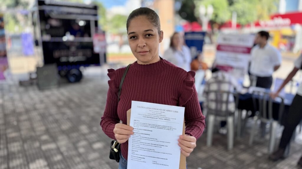 A imagem mostra uma mulher em um ambiente externo, provavelmente em um evento de empregabilidade ou feira de trabalho, segurando um currículo impresso voltado para a câmera. * A mulher, que está no centro da imagem, veste uma blusa de tricô cor de vinho com detalhes de babados nos ombros. Ela olha diretamente para a câmera com uma expressão serena. * Ela segura o currículo com as duas mãos, permitindo a leitura parcial dos dados, como o nome "Valéria Lúcia Gonçalves de Abreu Lima", além de seções como "Objetivo", "Experiência Profissional" e "Educação". * O cenário ao fundo é uma área aberta com piso de paralelepípedos. É possível observar pessoas ao fundo, mesas com materiais informativos, um veículo tipo "food truck" preto à esquerda e elementos que sugerem um evento público ou uma ação de recrutamento ao ar livre. A iluminação é de luz do dia, indicando um ambiente externo durante o período da manhã ou tarde.