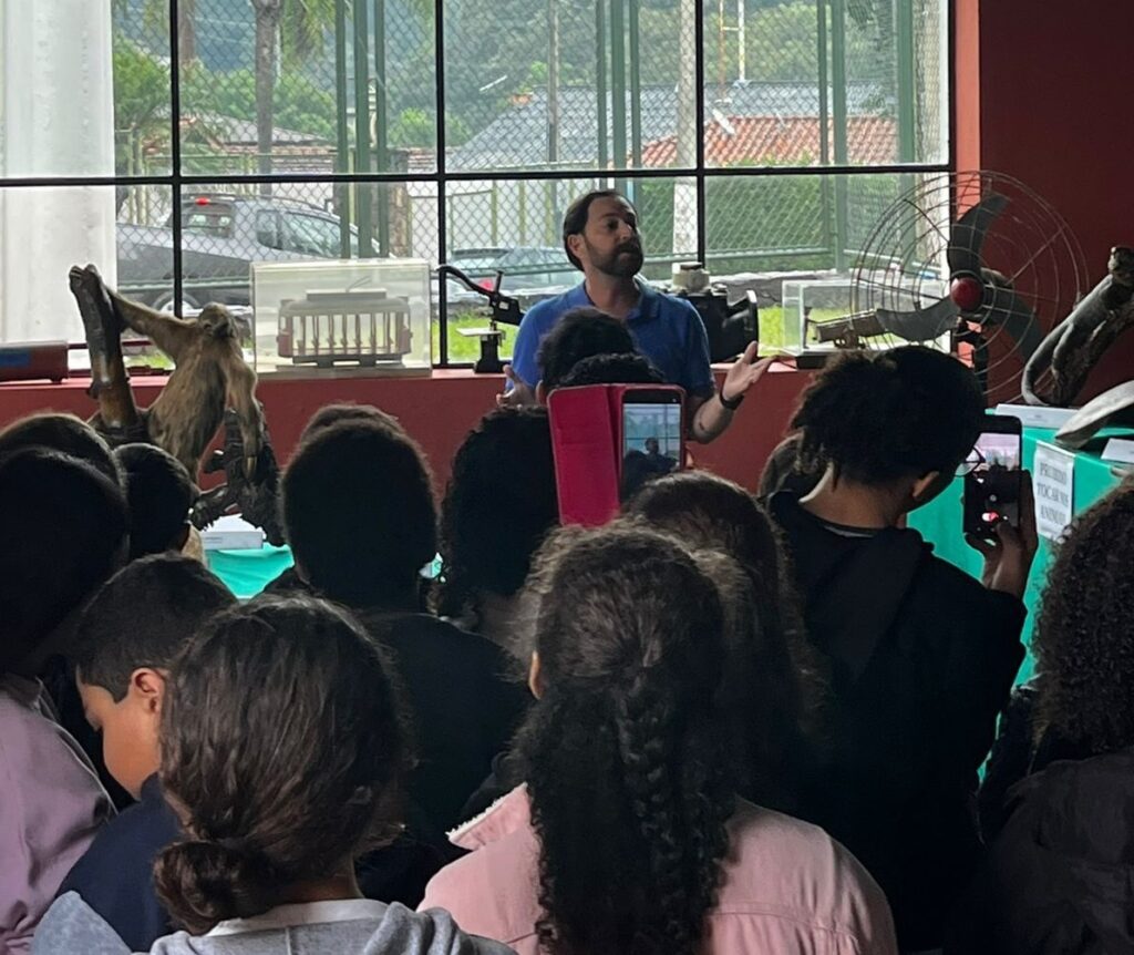 A imagem mostra uma cena educativa dentro de um museu ou centro histórico. Um homem, possivelmente o guia ou educador, está em pé na frente de um grupo de crianças e adolescentes, fazendo uma apresentação ou explicação. Ele veste uma camisa azul e está gesticulando com as mãos enquanto fala. As crianças estão de costas para a câmera, algumas segurando celulares para gravar ou tirar fotos. Atrás do homem, há uma grande janela com grades, que permite a entrada de luz natural e mostra uma área externa com cercas e edifícios. Na frente da janela, sobre uma prateleira ou bancada, estão expostos vários objetos, como um modelo de bonde, um ventilador antigo e o que parece ser um animal empalhado ou algum tipo de fóssil. Na parede à direita, há uma placa com o aviso em português: "PROIBIDO TOCAR NOS ANIMAIS", indicando que os visitantes não devem tocar nas peças expostas. A atmosfera da imagem sugere um ambiente de aprendizado e curiosidade, com as crianças atentas ao que está sendo apresentado. A iluminação natural destaca o educador e os objetos expostos, criando um contraste com o público.
