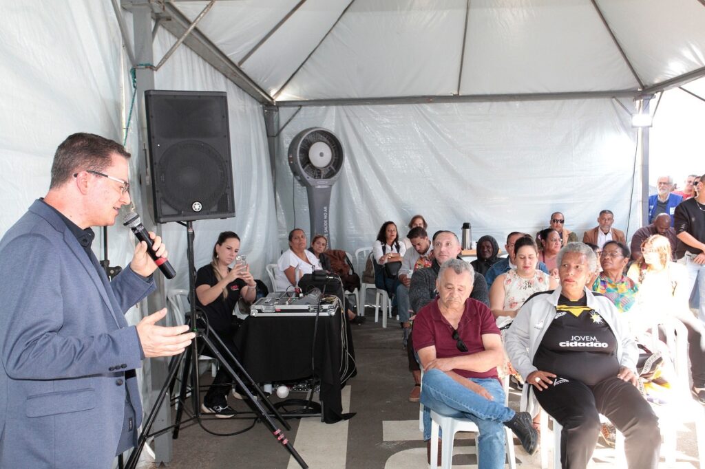 A imagem retrata um evento em um espaço coberto por uma tenda branca, onde um homem de terno azul, posicionado à esquerda, fala ao microfone para uma plateia sentada. * O palestrante, que usa óculos, está de perfil, gesticulando enquanto se dirige ao público. * À esquerda, próximo ao palestrante, uma mulher sentada manuseia um celular, com um equipamento de som (mesa de som e caixa acústica) logo atrás dela. * Ao fundo, um ventilador industrial de coluna está posicionado contra a lona lateral. * A plateia é composta por um grupo diversificado de pessoas sentadas em cadeiras de plástico brancas, prestando atenção à apresentação. * O ambiente possui iluminação natural vinda da parte frontal aberta da tenda e uma estrutura de metal que sustenta a cobertura.