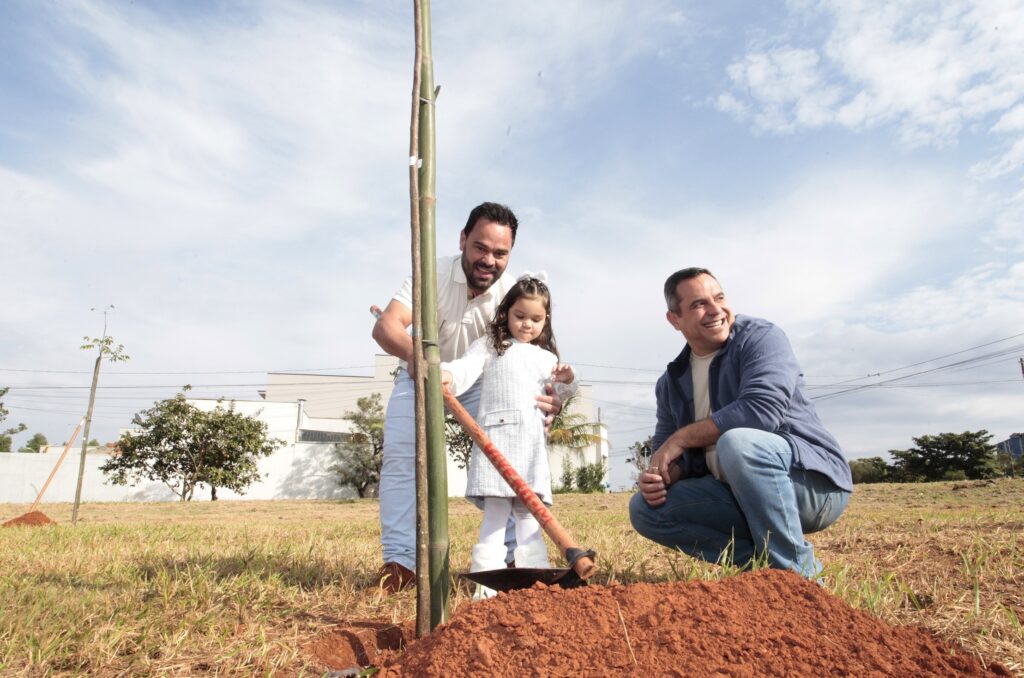 Nesta imagem, vemos um momento de plantio de uma muda de árvore ao ar livre. No centro da cena, um homem de camiseta polo branca e calça jeans clara está em pé, segurando uma pequena pá com a ajuda de uma menina pequena, que veste um casaco branco e botas peludas. Eles parecem estar cobrindo a raiz da muda, que está apoiada por uma estaca de bambu. À direita, outro homem, usando uma blusa azul sobre uma camiseta clara e calça jeans, está agachado no chão, sorrindo e olhando para o lado, observando a atividade. O cenário é um terreno aberto com grama seca e terra vermelha exposta no local do plantio. Ao fundo, sob um céu parcialmente nublado, vê-se uma construção branca e algumas árvores ao longe. A iluminação é de dia claro e ensolarado.
