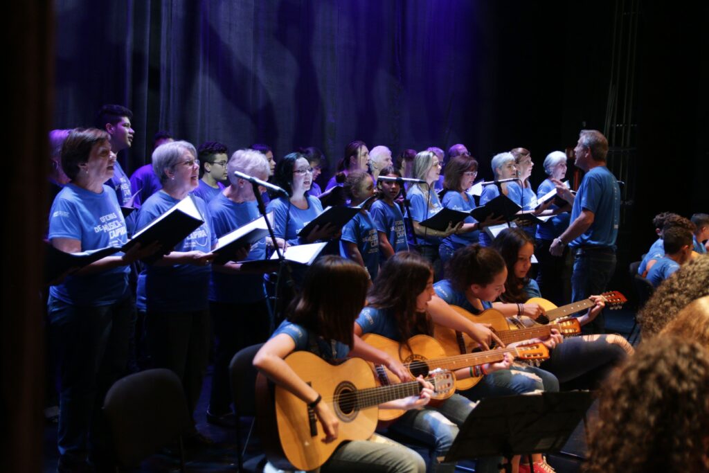 A imagem mostra uma apresentação musical em um palco com iluminação azul. Na frente, um grupo de jovens, principalmente meninas, está sentado tocando violões acústicos. Eles vestem camisetas azuis iguais. Atrás deles, um coral de pessoas de várias idades, também com camisetas azuis que têm a inscrição "OFICINAS DE MÚSICA CAIPIRA", está em pé cantando e segurando partituras. À direita, um maestro está de perfil, conduzindo o grupo com as mãos. O fundo é um palco escuro com cortinas pretas, destacando a iluminação azul que cria uma atmosfera focada e profissional. A cena transmite a ideia de uma apresentação organizada e harmoniosa, provavelmente de um grupo musical comunitário ou educacional.