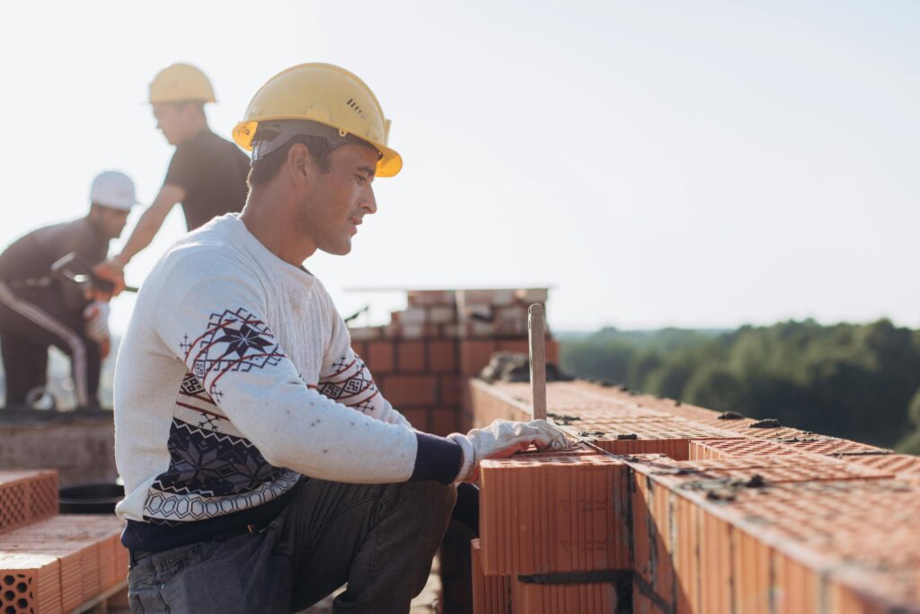 A imagem mostra uma cena de construção civil ao ar livre, em um dia ensolarado. Em primeiro plano, um trabalhador está agachado ao lado de uma parede de tijolos cerâmicos em fase de construção. Ele usa capacete de segurança amarelo, luvas de proteção e roupas de trabalho. Com uma das mãos apoiada sobre os tijolos, parece estar realizando o alinhamento ou acabamento da alvenaria. Ao fundo, outros dois trabalhadores também utilizam equipamentos de proteção individual (EPIs), como capacetes, e aparecem desfocados, sugerindo atividade contínua no canteiro de obras. A iluminação é natural e intensa, com o sol incidindo por trás dos trabalhadores, criando um efeito de contraluz. A profundidade de campo reduzida mantém o foco no trabalhador em primeiro plano, enquanto o restante da cena fica suavemente desfocado. A imagem transmite a ideia de trabalho em equipe, construção, desenvolvimento urbano e atividade profissional na área da construção civil.