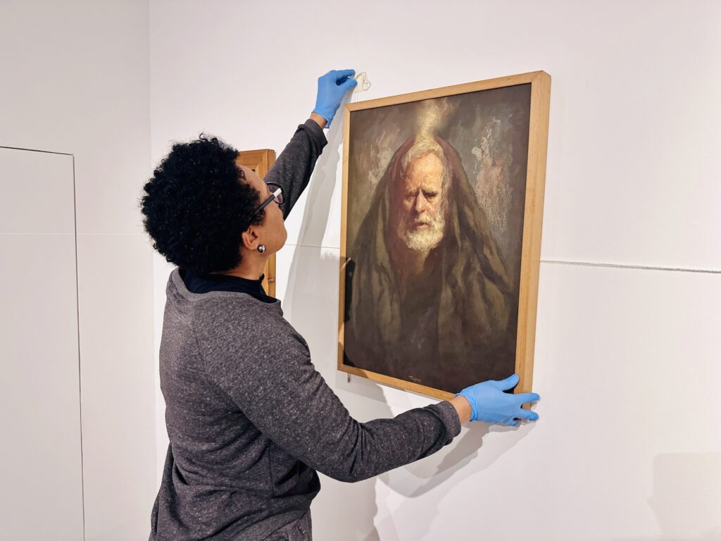 Esta imagem mostra uma pessoa de perfil, com cabelos curtos e escuros, vestindo um moletom cinza e usando luvas de proteção azuis, enquanto instala ou ajusta um quadro em uma parede branca. O quadro, que tem uma moldura de madeira clara, retrata um homem idoso com barba branca e um capuz ou manto escuro sobre a cabeça, com um estilo artístico clássico e tons terrosos. A pessoa segura o quadro com as duas mãos, posicionando-o com cuidado. A cena parece acontecer em uma galeria ou museu, dado o cuidado com o manuseio da obra de arte.