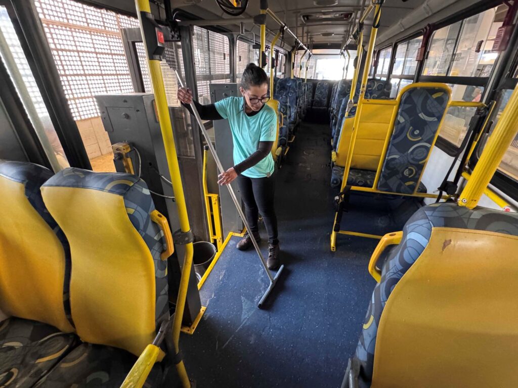 A imagem mostra o interior de um ônibus urbano sendo limpo. Aqui estão os detalhes: A pessoa: Uma mulher usando óculos, camiseta azul-clara e calça preta está passando um rodo no piso do ônibus. Ela parece concentrada na tarefa. O interior: O ônibus tem um corredor central com bancos de estofamento amarelo e padrões geométricos em tons de azul e cinza. Os suportes e barras de apoio são de cor amarela vibrante. O ambiente: O veículo está vazio, sem passageiros, indicando que a limpeza provavelmente ocorre entre viagens ou no final de uma rota. Através das janelas laterais, é possível ver o ambiente de um terminal de transporte, com estruturas metálicas e paredes de blocos de concreto.
