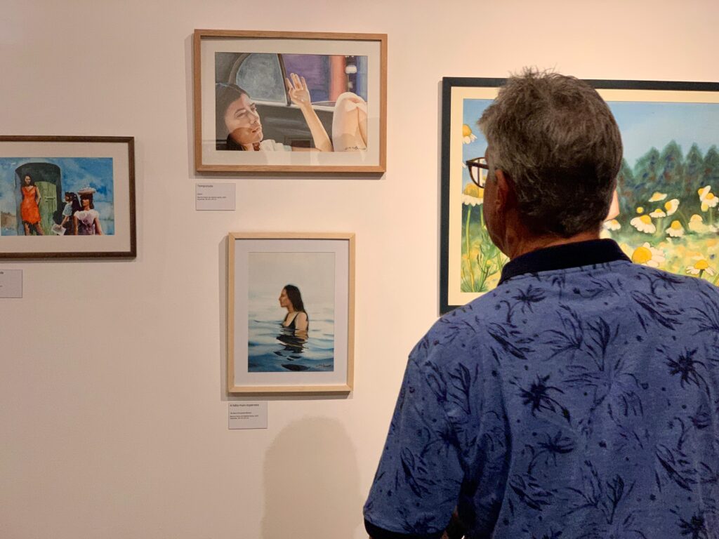Uma cena em uma galeria ou exposição de arte. De costas, vemos um homem vestindo uma camisa azul estampada e óculos, observando atentamente três quadros pendurados em uma parede branca.
O quadro superior esquerdo: Retrata uma mulher no banco de trás de um carro, olhando para fora com uma das mãos levantadas. A legenda abaixo diz "Temporada".
O quadro inferior esquerdo: Mostra uma mulher mergulhada na água, vista de perfil. A legenda diz "A falta mais esperada".
O quadro à esquerda (mais afastado): Exibe uma cena externa com três mulheres ao ar livre.
À direita: Há um quadro maior, parcialmente visível, que parece retratar um campo de flores, possivelmente margaridas.
É um ambiente bem iluminado, com molduras de madeira clara e preta, criando uma atmosfera contemplativa.