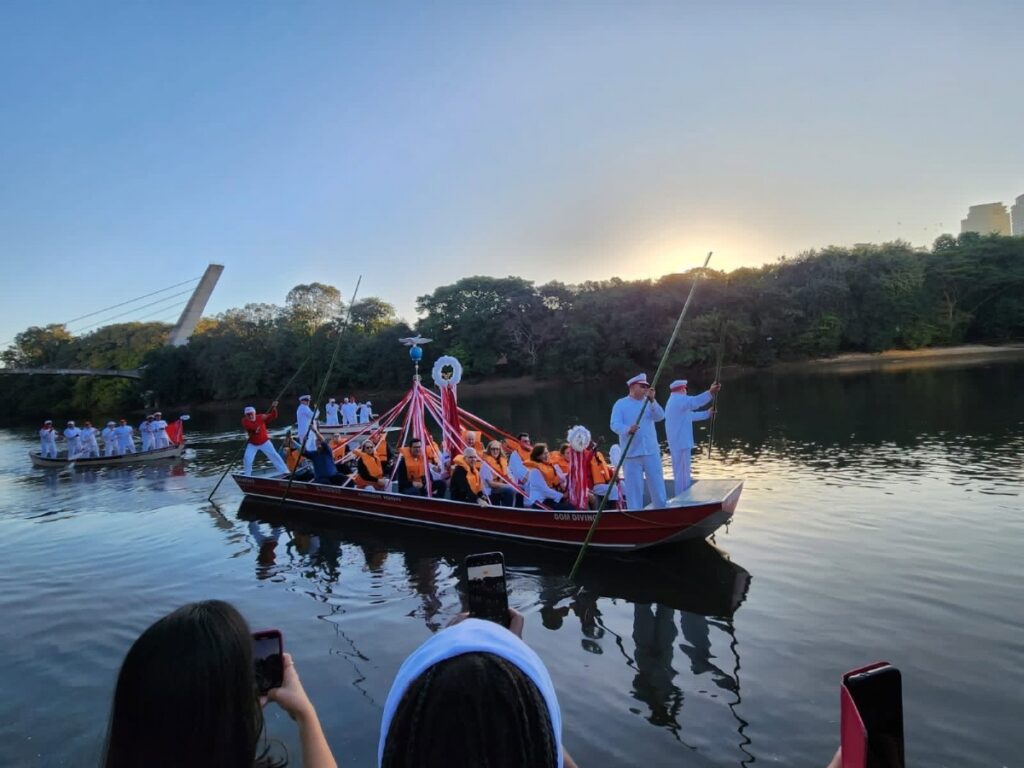 Foto de um cortejo ou procissão sobre o rio ao pôr do sol. Principais elementos: Cenas principais: duas canoas grandes ocupadas por muitas pessoas; a canoa da frente está mais próxima e tem enfeites centrais com fitas vermelhas e brancas formando um mastro decorativo. Pessoas: muitos ocupantes vestidos majoritariamente de branco; vários usam coletes salva-vidas laranja. Alguns tripulantes em pé manuseiam longas varas para guiar as embarcações. Ação e atmosfera: sensação festiva e cerimonial — as fitas, coroas decorativas e a postura formal de alguns (chapéus brancos) sugerem evento religioso ou cultural. Luz suave do pôr do sol reforça clima tranquilo e solene. Ambiente: rio calmo com reflexos nítidos da canoa e das pessoas na água; margem arborizada ao fundo; à esquerda nota-se uma ponte moderna parcialmente visível; céu claro e gradiente de cores por causa do entardecer. Primeiro plano: espectadores na margem filmando/registrando com celulares — parte das cabeças e celulares aparecem na base da foto. Detalhes menores: uma segunda canoa ao fundo com mais participantes, e algumas aves no céu distante.