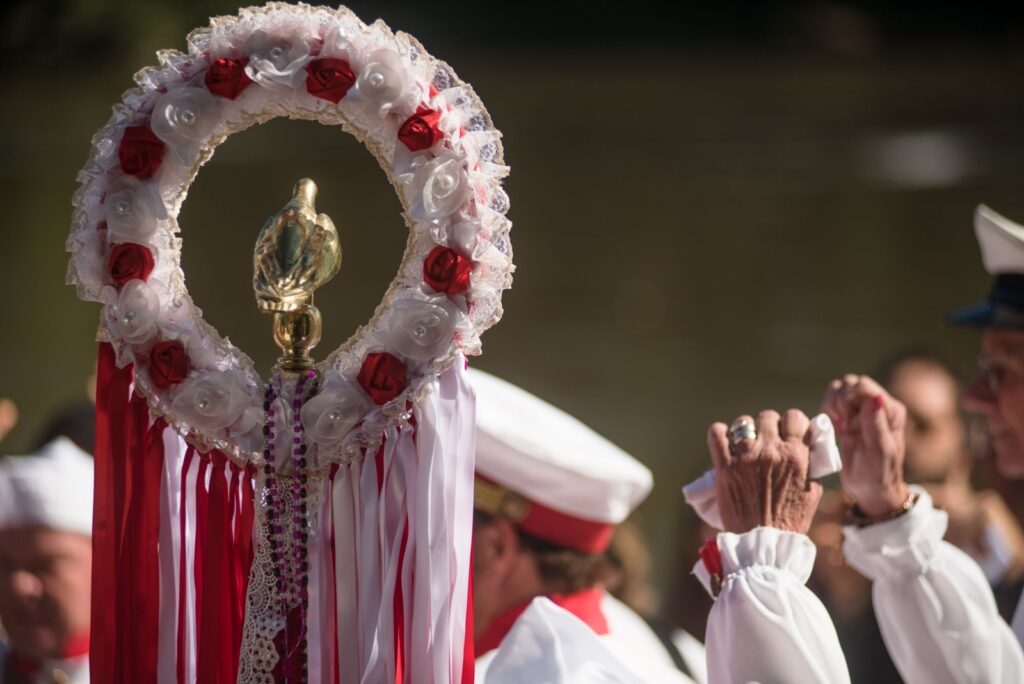 Foto de uma procissão cerimonial, com foco em um estandarte enfeitado e participantes em trajes brancos. Principais elementos: Estandarte central: arco circular decorado com renda e flores artificiais brancas e vermelhas, pé metálico dourado com detalhe esculpido no topo. Longas fitas vermelhas e brancas pendem abaixo, junto a rendas e colares de contas. Pessoas: à direita aparecem mãos de pessoas idosas em gesto de oração ou saudação — punhos cerrados e dedos entrelaçados, com alguns anéis e um lenço branco. Ao fundo vêem-se figuras com boinas/brancos e detalhes vermelhos no uniforme. Cores e luz: predominância de branco e vermelho no primeiro plano; iluminação quente e suave, com fundo desfocado em tons terrosos (possivelmente água). Ambiente e atmosfera: sensação religiosa, festiva e solene — pode ser parte de uma procissão marítima ou celebração tradicional. O foco no estandarte e nas mãos transmite devoção e ritual. Detalhes menores: fundo fora de foco que sugere um rio ou lago; não é possível identificar rostos ou nomes das pessoas.