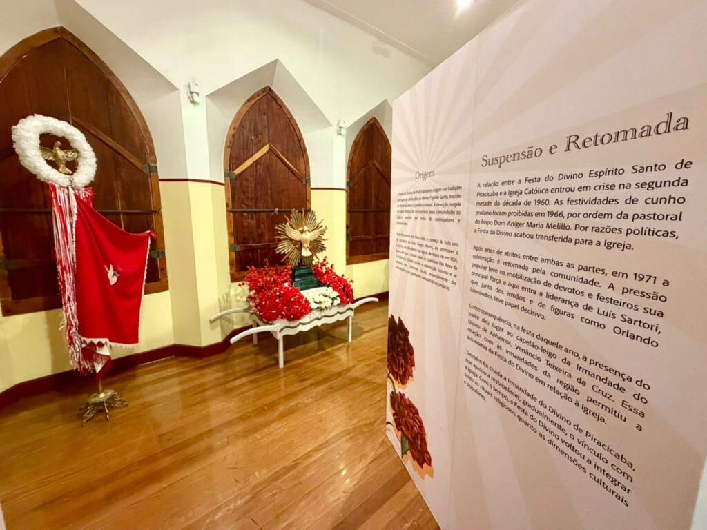 A imagem mostra parte da exposição "200 Anos de Festa do Divino Espírito Santo de Piracicaba", dentro de um espaço com arquitetura histórica.Detalhes da cena:Ambiente: Sala com piso de madeira, paredes brancas com faixa amarela na base e janelas/portas em arco de madeira escura. O estilo lembra uma capela ou prédio antigo.Objetos expostos:À esquerda: Um mastro ou bandeira do Divino. Tem um pano vermelho com a pomba branca bordada, franjas brancas e vermelhas, e no topo uma coroa branca com a pomba dourada no centro.Ao centro: Uma pequena carroça branca decorada com flores vermelhas e brancas. Em cima dela está o símbolo da pomba do Divino dentro de um resplendor dourado, típico das coroas usadas na festa.Painel informativo: À direita, um painel grande com fundo claro. O título em destaque é "Suspensão e Retomada". O texto conta que a festa foi proibida em 1966 por ordem do bispo Dom Aniger Maria Melillo, e retomada em 1971 pela pressão da comunidade e liderança de figuras como Luís Sartori, Orlando Louvandini e o padre Venâncio Teixeira da Cruz. Também fala da criação da Irmandade do Divino de Piracicaba nesse período. O painel tem ilustrações de flores vermelhas na base.Clima: A exposição mistura objetos rituais, história e memória. As cores vermelho e branco predominam, remetendo às cores tradicionais da Festa do Divino.É um registro que mostra tanto a religiosidade quanto a resistência cultural da festa mais antiga de Piracicaba.