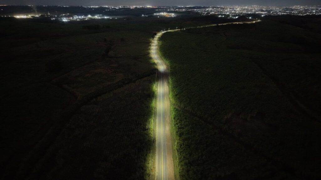 Esta é uma vista aérea noturna de uma estrada sinuosa que atravessa uma área rural.
Estrada: A via é o ponto central da imagem, iluminada artificialmente por postes ao longo de toda a sua extensão, o que cria um contraste marcante com a escuridão ao redor. A iluminação destaca o traçado da estrada, que se curva suavemente pelo terreno.
Ambiente: A estrada é ladeada por densa vegetação ou plantações escuras, que ocupam a maior parte do campo de visão, enfatizando o isolamento do local.
Fundo: Ao longe, no horizonte, vê-se o brilho das luzes de uma área urbana ou cidade, criando um contraste entre a calmaria do campo iluminado e a agitação das luzes distantes. A cena transmite uma sensação de tranquilidade e vastidão noturna.