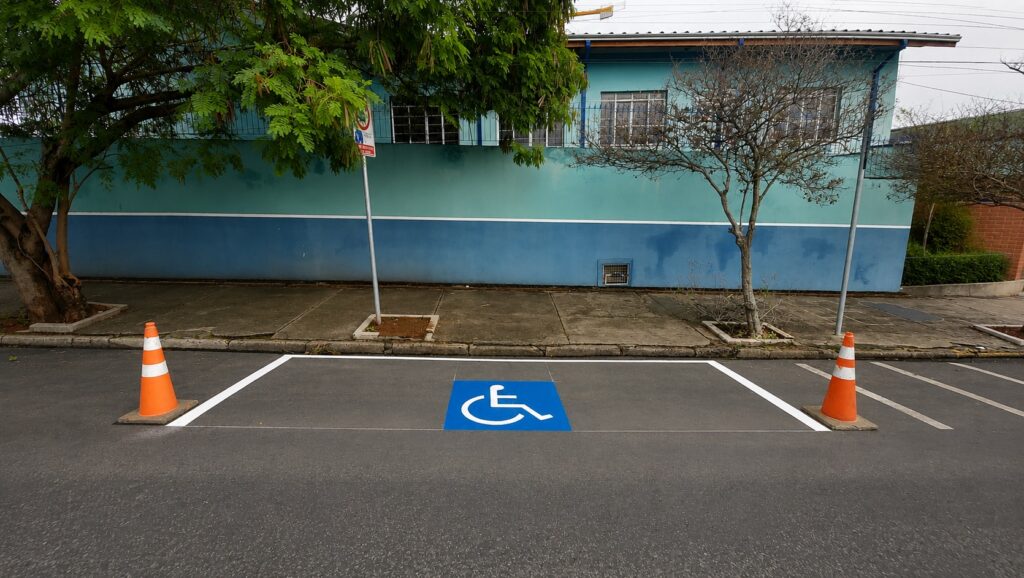 Vaga de estacionamento destinada a pessoas com deficiência, recém-pintada. Elemento central: símbolo internacional de acessibilidade pintado em azul no centro da vaga. Marcação: faixas brancas delimitando a vaga e linha de bordo; pintura parece recente. Sinalização física: dois cones laranja (um em cada extremidade frontal) bloqueiam temporariamente a vaga. Ambiente: rua asfaltada sem veículos estacionados; meio-fio e calçada de concreto logo atrás. Plano de fundo: prédio baixo pintado em tons de turquesa/azul com janelas gradeadas; duas árvores pequenas junto à calçada (uma à esquerda com copa densa, outra à direita mais rala). Observações: poste de sinalização na calçada (sinal não totalmente legível na foto) e áreas de jardineira ao redor das árvores.