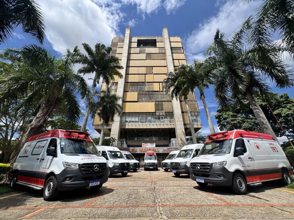 Esta imagem mostra um grupo de ambulâncias do SAMU (Serviço de Atendimento Móvel de Urgência) alinhadas em frente a um prédio alto de arquitetura moderna, que é a Prefeitura Municipal. O cenário é um dia ensolarado com céu azul e algumas nuvens. As ambulâncias são brancas com detalhes em vermelho, todas com a marca do SAMU 192 visível. Ao fundo, o prédio imponente apresenta um design com elementos de concreto e painéis verticais.