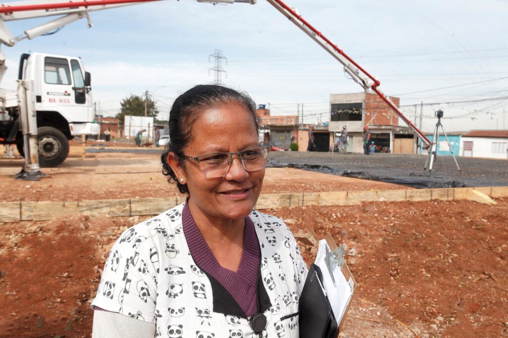 Uma mulher em primeiro plano ocupando a metade direita da imagem, vista da altura do peito para cima. Detalhes visíveis: Aparência e roupa: mulher de meia-idade com cabelo escuro preso para trás, pele morena clara, usa óculos de grau e brincos pequenos. Veste uma blusa/camisa com estampa de pandas em tons branco/preto e suéter roxo por baixo; segura prancheta e papéis na mão esquerda. Expressão e postura: sorriso leve e expressão tranquila; postura relaxada, levemente virada para a câmera. Cena de fundo: canteiro de obras urbano em andamento. No centro e à esquerda aparecem um caminhão-bomba branco e o braço/tubo vermelho de bombeamento de concreto em operação, despejando material sobre uma área coberta por lona preta e malha de vergalhões. Pessoas e atividade: vários trabalhadores ao fundo, alguns com capacetes e EPIs, ocupados na concretagem e no nivelamento do piso; um tripé com equipamento topográfico está posicionado à direita. Ambiente e construções: solo de terra avermelhada no primeiro plano; fôrmas de madeira delimitando a área concretada. Ao fundo, casas e pequenos prédios de alvenaria, fiação aérea e postes. Iluminação e clima: dia claro, luz solar difusa sem sombras duras — aparência de manhã ou tarde ensolarada.