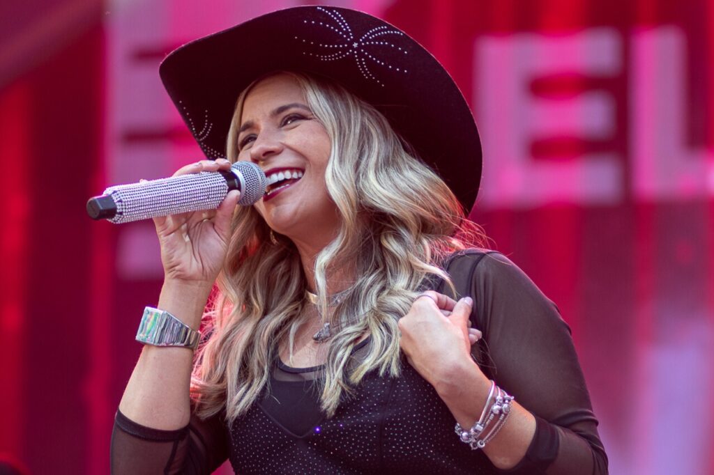 Mulher sorrindo enquanto canta em um show ao vivo, em close médio. Detalhes observáveis: Expressão e pose: sorriso largo e natural, olhando ligeiramente para a esquerda; segura o microfone próximo à boca com a mão direita e com a esquerda toca o peito, num gesto caloroso e envolvente. Rosto e cabelo: pele de tonalidade clara, cabelos loiros com mechas mais claras, ondulados e soltos até os ombros. Roupa e acessórios: usa um chapéu estilo cowboy preto com detalhes brilhantes; blusa preta com mangas transparentes e corpete pontilhado de pequenos brilhos; colar delicado; relógio prateado largo no pulso esquerdo e várias pulseiras prateadas no pulso direito. Microfone: revestido de strass ou cristais brilhantes, destacando-se como acessório glamouroso. Iluminação e cenário: luzes de palco quentes (tons rosados/vermelhos) criando um fundo desfocado colorido; atmosfera claramente de show ao vivo. Tom geral: imagem transmite energia positiva, carisma e conexão com o público — sensação de performance animada e confiante.