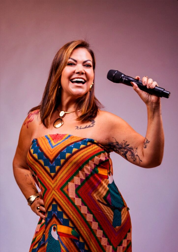 Esta foto mostra uma mulher sorridente e cheia de energia, segurando um microfone. Aqui estão os detalhes: A Pessoa: Ela tem cabelos castanhos na altura dos ombros, uma expressão de muita alegria e está posando para a câmera. Ela usa um colar dourado com um pingente grande e uma pulseira brilhante. É possível notar algumas tatuagens em seus braços e colo. A Roupa: Ela veste um vestido tomara-que-caia colorido, com uma estampa vibrante de padrões geométricos (em tons de laranja, amarelo, verde e azul) e desenhos de tucanos na parte inferior. A Pose: Com a mão direita na cintura e a esquerda levantada segurando um microfone preto, ela parece estar se apresentando ou se divertindo em um momento de entretenimento. O Estilo: O fundo é liso, em um tom neutro rosado/lilás, o que destaca bastante as cores vibrantes do vestido e a expressão alegre da mulher.