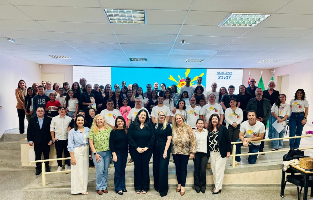 Foto de um grande grupo posando em um auditório/plateia para uma foto oficial. Principais pontos: Ambiente: sala com teto rebaixado e painéis de iluminação, palco com escadas laterais e uma grande tela LED ao fundo (na tela aparece a data 30-06-2026 e o horário 21:07). Pessoas: cerca de 60–70 pessoas de idades variadas (crianças, adolescentes, adultos e idosos), dispostas em fileiras sobre o palco e nas escadas — muitas delas sorrindo. Roupa e identidade visual: vários participantes usam camisetas brancas com um logo colorido (parece desenho/mascote e a palavra “FAM”/algo parecido), outros vestidos de forma mais formal ou casual; há uniformidade parcial que sugere um evento ou projeto comunitário/escolar. Acessórios e objetos: algumas pessoas seguram certificados ou papéis; há bandeiras ao fundo (provavelmente institucional/municipal/estadual) à direita. Atmosfera: sensação de celebração, conclusão de projeto ou cerimônia (entrega de certificados, formatura ou confraternização). Detalhes do local: corrimão amarelo na frente, piso em granito, cadeiras e mesas laterais visíveis.