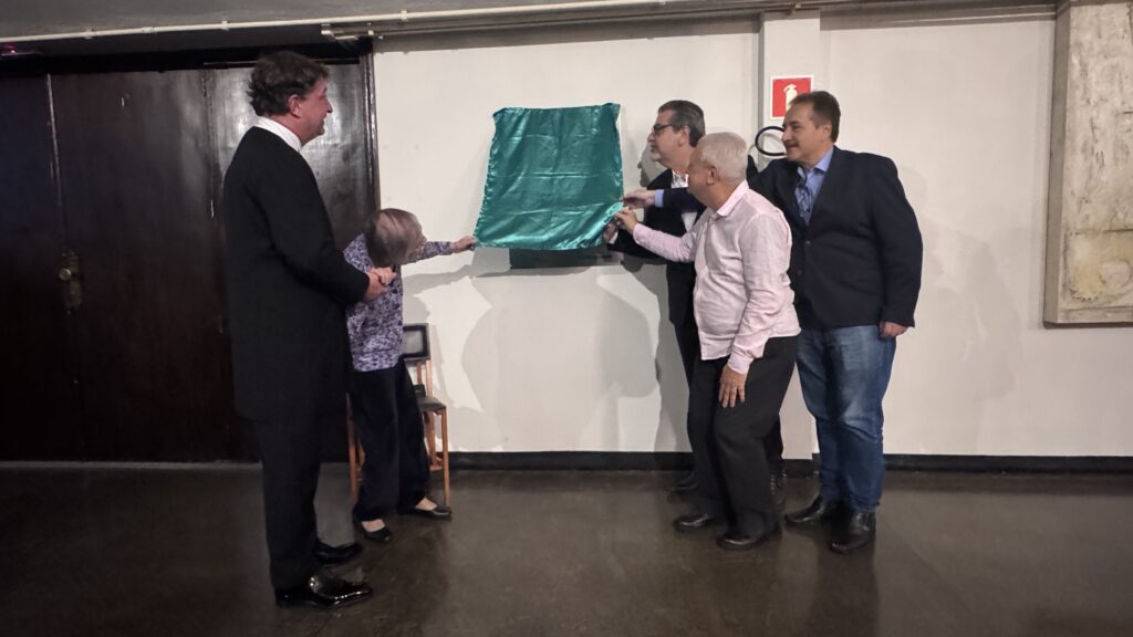 Nesta imagem, cinco pessoas estão reunidas em um ambiente interno, aparentemente participando de uma cerimônia de descerramento de uma placa. À esquerda, um homem de terno preto está segurando o braço de uma senhora idosa com blusa estampada, que está ajudando a puxar o tecido. À direita, um grupo de três homens está posicionado junto à parede. Um deles, vestindo camisa rosa clara e calça escura, segura a ponta do tecido verde que cobre a placa, enquanto os outros dois — um de terno escuro e camisa branca, e outro de blazer escuro, camisa azul e jeans — observam o momento. Eles parecem estar no processo de revelar o que está atrás do tecido, que está fixado em uma parede branca. Há uma porta de madeira escura à esquerda e uma sinalização de extintor de incêndio na parede ao fundo.