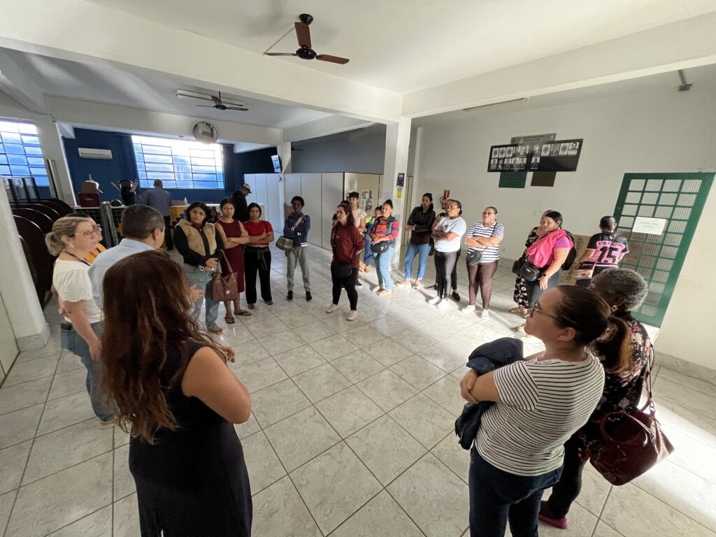 Uma sala comunitária ou hall amplo com piso de cerâmica claro onde um grupo de cerca de 20 pessoas, majoritariamente mulheres, está reunido formando um semicírculo/roda improvisada. Parecem ouvir alguém que está à esquerda (fora do centro da foto) — várias pessoas de braços cruzados, outras com bolsas, algumas em postura atenta. Detalhes visuais: Ambiente: teto branco com ventiladores de teto, paredes claras; janelas grandes com grades e vidro azul ao fundo, iluminação natural forte entrando pela esquerda. Piso: azulejos quadrados claros com rejunte visível, refletindo a luz. Pessoas: diversidade de idades; roupas casuais (camisetas, blusas, jaquetas, calças e vestidos). Algumas usam cores vivas (rosa, listras, vermelho), outras tons neutros. Há também quem use óculos e acessórios como bolsas e anéis. Movimento/comportamento: maioria em posição de espera/escuta, poucas conversando entre si; expressão geral séria ou concentrada. Mobiliário e itens: ao fundo, à esquerda, há móveis empilhados e um balcão; à direita, quadro de avisos/placar verde inclinado na parede; fotos ou quadro de homenagens na parede direita. Composição fotográfica: imagem feita em ângulo levemente superior e ampla (lente grande-angular), causando leve distorção nas bordas e mostrando bastante do piso e do contexto da sala.