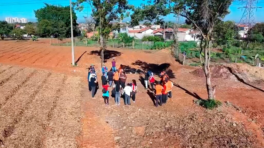 Uma área de terra arada ou terreno aberto, vista de cima em ângulo — provável horta/terreno comunitário — onde um grupo de cerca de 20 pessoas está reunido formando pequenos aglomerados no centro da cena. Grupo: aproximadamente 18–22 pessoas, algumas em pares e outras em pequenos círculos; várias usam coletes laranja (possivelmente equipe/voluntários) e há roupas coloridas (azul, vermelho, branco). A maioria está de pé, conversando ou olhando para o chão; não há rostos identificáveis à distância. Terreno e solo: solo avermelhado e seco, com partes revolvidas/aratadas à esquerda e pequenos montes de terra e palha; faixa central mais compacta onde o grupo está em pé. Vegetação e árvores: duas árvores de médio porte próximas ao grupo (uma mais central e outra à direita), projetando sombras longas no solo; ao fundo pequenas áreas cultivadas/plantio e vegetação mais densa. Infraestrutura: poste de iluminação/energia próximo ao centro-esquerdo; torres e cabos de alta tensão visíveis à direita e mais ao fundo; residências com telhados vermelhos e alguns prédios baixos na linha do horizonte. Céu e iluminação: céu claro e azul, luz solar forte vindo de cima-direita (sombras definidas), sugerindo manhã ou início da tarde. Impressão geral: reunião de campo ou atividade comunitária/voluntária num espaço de cultivo urbano/periurbano — cena transmite organização em ambiente rural dentro de contexto urbano próximo.