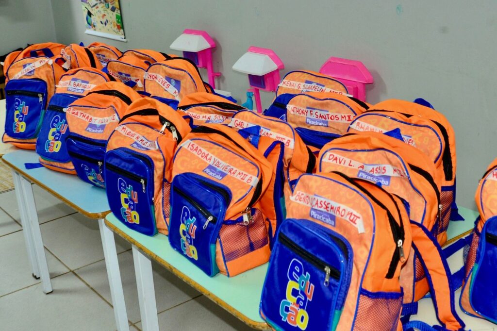 Mesa com várias mochilas escolares alinhadas em fileiras, provavelmente preparadas para entrega. Mochilas: predominância de laranja com bolsos frontais azuis; logo colorido com a palavra que remete a "educação" visível nos bolsos. Etiquetas: em cada mochila há uma etiqueta branca com nomes escritos à mão em caneta vermelha/laranja (nomes de alunos e turma/identificação). Quantidade e arranjo: dezenas de mochilas dispostas lado a lado sobre duas mesas compridas, formando três fileiras visíveis na imagem. Ambiente: sala com piso de cerâmica clara e parede cinza ao fundo; ao fundo também há pequenos suportes/objetos rosa (possivelmente luminárias ou peças infantis) encostados na parede. Estado e propósito: aparência organizada, sugere preparação para distribuição escolar (kits ou material para alunos).