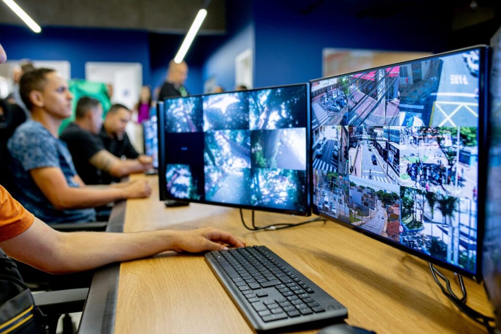 A imagem mostra uma sala de monitoramento com várias pessoas sentadas diante de computadores. Nos dois monitores principais, há diversas câmeras de vigilância exibindo cenas de ruas, cruzamentos e áreas urbanas — indicando um sistema de controle de segurança ou trânsito. O ambiente tem paredes azuis, iluminação moderna e um ar de centro operacional tecnológico, com foco em observação e análise em tempo real.