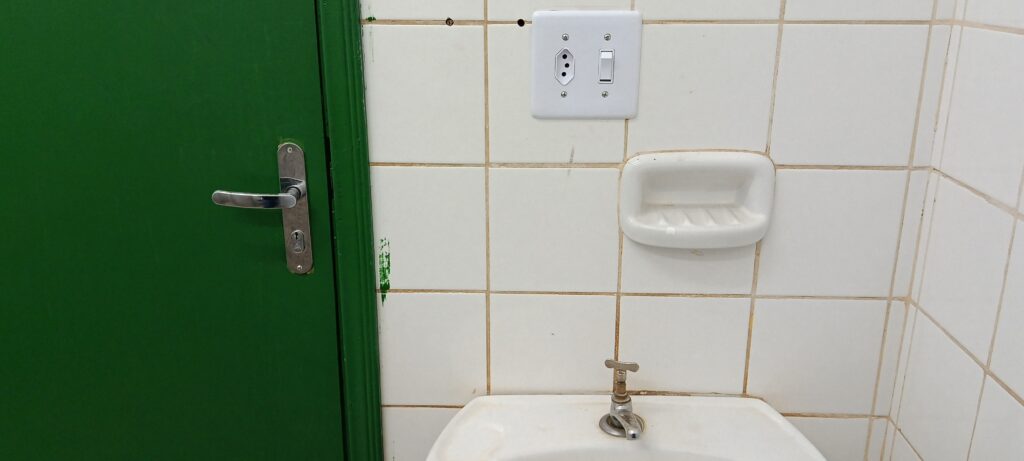 A imagem mostra um detalhe de um banheiro ou área de lavatório. À esquerda: Há uma porta verde vibrante com uma maçaneta de metal prateado. É possível notar uma pequena marca de tinta verde na parede branca logo ao lado da batente da porta. Parede: A parede é revestida com azulejos quadrados brancos. Elementos na parede: Acima, há uma tomada e um interruptor de luz brancos instalados na parede. Um pouco mais abaixo, há uma saboneteira de cerâmica branca embutida ou fixada na parede. Base: Na parte inferior, vê-se a borda de uma pia (lavatório) de cerâmica branca, com uma torneira de metal simples instalada no centro. O ambiente parece funcional e simples.