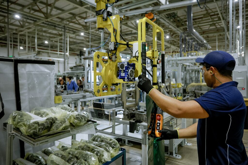Foto de um ambiente industrial/linha de montagem com um trabalhador operando um manipulador mecânico. Cena geral: interior de fábrica amplo, bem iluminado, com estrutura metálica do teto e várias máquinas e esteiras ao fundo. Pessoa: trabalhador de perfil direito, usando boné azul, óculos de proteção, protetores auriculares e luvas pretas — segurando o painel de controle do equipamento. Equipamento: um braço/manipulador industrial amarelo suspenso com garras ou gancho, posicionado sobre uma esteira ou bancada; painel de controle laranja com botões visíveis na mão do operador. Materiais: prateleiras na frente esquerda com peças embaladas em plástico transparente (parecem componentes metálicos ou eixos). Atividade: parece-se com operação de movimentação/posicionamento de peças na linha de montagem — o operador guia o equipamento para pegar ou depositar componentes. Ambiente e segurança: área organizada, uso de EPI pelo trabalhador, tubulações e equipamentos industriais ao fundo, sugerindo processo automatizado ou semi-automatizado.