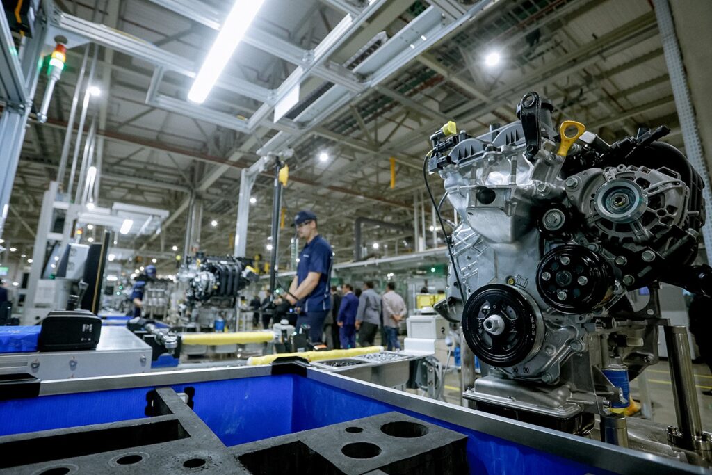 Esta imagem mostra o interior de uma linha de montagem industrial, especificamente voltada para a fabricação de motores de automóveis. Destaque em primeiro plano: À direita, vemos um motor de combustão interna completo e montado, exibindo polias, correias, alternador e outras peças metálicas, montado em um suporte de transporte. Ambiente de Produção: O cenário é uma fábrica moderna, com teto alto, estruturas metálicas expostas e iluminação clara e difusa. Vê-se uma linha de montagem automatizada ou semi-automatizada, com caixas plásticas azuis de armazenamento em primeiro plano, contendo componentes ou ferramentas em organizadores de espuma preta. Trabalhadores e Visita: Ao fundo, um operário uniformizado (camiseta azul e boné) trabalha ativamente na linha. Atrás dele, há um grupo de pessoas vestidas em trajes sociais ou casuais, sugerindo uma visita guiada ou um tour corporativo pela fábrica, observando o processo de produção.