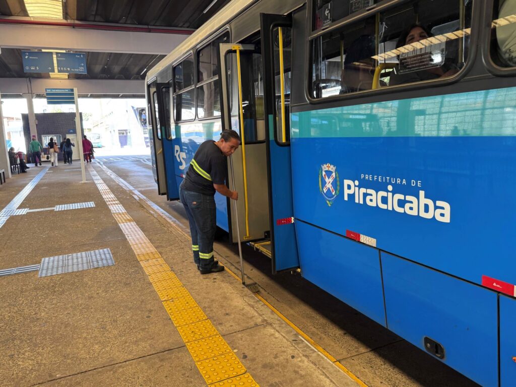 Esta foto mostra uma cena cotidiana no terminal de ônibus de Piracicaba. Aqui estão os detalhes principais: Ação: Uma funcionária da limpeza está varrendo a plataforma, exatamente na borda onde o ônibus encosta, enquanto o veículo está parado com a porta aberta. A Funcionária: Ela está vestindo um uniforme de trabalho escuro (camiseta e calça) com faixas reflexivas nas pernas e mangas, indicando segurança. O Ônibus: É um ônibus urbano azul, com o brasão da "Prefeitura de Piracicaba" estampado na lateral. A porta do meio está aberta para o embarque/desembarque. O Ambiente: O terminal é coberto, com piso de concreto que possui faixas podotáteis amarelas para acessibilidade. Ao fundo, pode-se ver placas de sinalização suspensas (indicando "Bilheteria", "Sanitários", etc.) e algumas pessoas caminhando pela plataforma, o que indica que o terminal está em funcionamento.