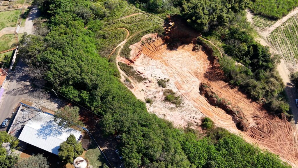 Vista aérea de uma área de escavação cercada por vegetação densa. Em destaque, uma grande cava de solo avermelhado com marcas visíveis de esteiras de maquinário formando sulcos concêntricos e radiais; o fundo da cava é mais claro, com partes de terra batida e manchas de vegetação residual. No topo direito da cava há uma escavadeira/amarelada estacionada junto à borda, parcialmente sombreada por árvores. Ao redor da área escavada há mosaico de vegetação: à esquerda e embaixo abundante copa de árvores verdes forming um cinturão contínuo; à direita e ao fundo aparecem trechos de pasto/cultivo e caminhos de terra que delimitam lotes. No canto inferior esquerdo vê‑se um conjunto de construções com telhado branco, uma rua asfaltada com postes e algumas árvores cujas sombras projetam ramos sobre o solo. A iluminação é solar, com sombras nítidas, indicando dia claro. A cena sugere atividade recente de terraplenagem ou extração num entorno semiurbano/rural.