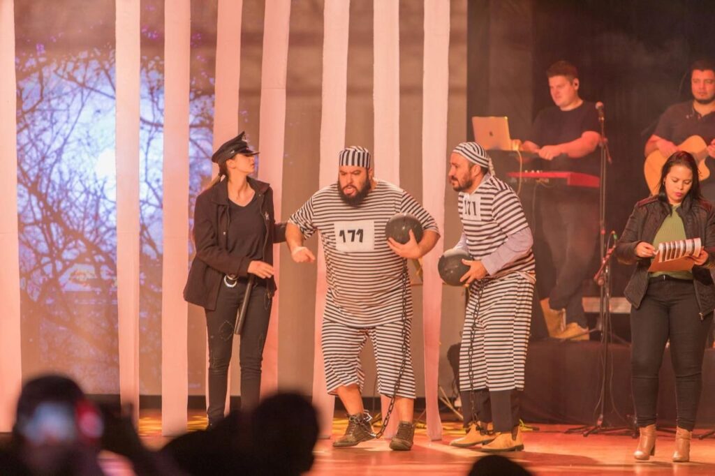 Esta é uma foto de uma cena teatral ou musical, muito bem produzida e com tom humorístico! Personagens: Há três personagens principais no palco: À esquerda, uma pessoa vestida de policial, com boné, jaqueta preta e segurando um cassetete, interagindo com os outros. Ao centro e à direita, dois homens vestidos como prisioneiros de desenho animado clássico (uniforme de listras horizontais pretas e brancas e gorro combinando). Eles têm o número "171" no peito, seguram bolas de ferro (cenográficas) e têm correntes presas aos pés. Músicos: Ao fundo, um pouco fora de foco, há um músico tocando teclado e outro tocando violão, sugerindo que é uma apresentação musical encenada. Outra Participante: No canto inferior direito, há uma mulher vestindo uma blusa verde, jaqueta preta e calça jeans, segurando uma prancheta, parecendo parte da equipe de produção ou uma personagem administrativa na cena. Cenário: O fundo é composto por grandes faixas verticais brancas (que lembram as grades de uma cela) e uma projeção de árvores secas ou galhos. Atmosfera: A iluminação é quente e o ambiente parece ser um teatro ou auditório. A expressão do prisioneiro ao centro, com uma cara de bravo ou surpreso, dá um toque muito engraçado à performance.