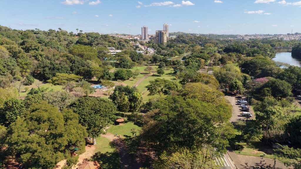 Uma vista panorâmica de um grande parque urbano arborizado com um rio à direita e prédios ao fundo, em dia claro e ensolarado. Panorama geral: cena ampla em perspectiva aérea/alto — muito verde contínuo com árvores densas, clareiras gramadas e caminhos que cortam o parque; rio sereno corre pela margem direita até um pontilhão ao fundo; céu azul com poucas nuvens. Vegetação e áreas abertas: mistura de copas altas e áreas de gramado; várias árvores de diferentes tamanhos e tonalidades de verde; pequenas clareiras com bancos e quiosques com telhado de cerâmica visível no primeiro plano inferior. Caminhos e infraestrutura: trilhas e caminhos de terra e pavimentados atravessam o parque em diversas direções; uma área circular com fonte ou espelho d’água de cor azul clara aparece mais ao centro-esquerdo; postes de iluminação e postes de sinalização espalhados. Uso e circulação: estacionamento com carros alinhados na marge direita-central; algumas pessoas pequenas vistas caminhando nos caminhos (distantes); estrada/ciclovia no canto inferior direito com faixas de pedestres. Contexto urbano: ao fundo, além do cinturão verde, surgem casas e prédios mais altos (dois edifícios altos bem visíveis no centro), indicando transição para área urbana residencial. Colinas com mais vegetação ao longe. Iluminação e clima: luz solar forte e direta, sombras definidas das árvores sobre o gramado; sensação de dia seco e limpo, provável clima de estação seca ou outono/inverno seco. Atmosfera: ambiente sereno e bem conservado, mistura equilibrada de natureza e infraestrutura urbana — parece um parque público amplo, ideal para caminhadas, lazer e atividades ao ar livre.
