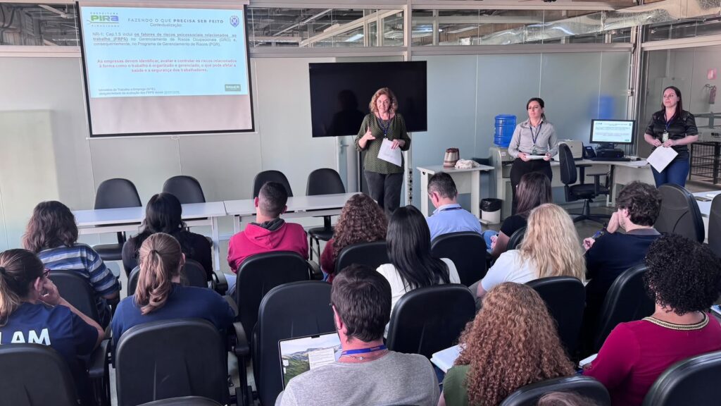 Foto de uma sessão de apresentação/treinamento em sala de reuniões. Cena geral: fileira de cadeiras ocupadas por participantes vistos de costas, olhando para a frente onde três apresentadoras ficam em pé. Apresentadoras: três mulheres na frente — uma central falando e gesticulando enquanto segura folhas; as outras duas, à direita, estão em posição de apoio com papéis e pranchetas. Público: cerca de 15–20 pessoas sentadas em cadeiras pretas, algumas anotando em cadernos ou usando laptop; há diversidade de idades e estilos de roupa (casual a profissional). Recursos visuais e equipamentos: projetor com tela à esquerda exibindo slide (logo no canto superior esquerdo e texto sobre contexto/NRs visíveis), grande monitor/TV central desligado ou sem conteúdo; computador e mesa de apoio à direita. Ambiente e mobiliário: sala clara com divisórias de vidro ao fundo, teto com dutos aparente; garrafa d'água/galão em suporte; mesas brancas e cadeiras pretas de escritório. Atmosfera: ambiente formal e concentrado, típico de capacitação institucional ou reunião técnica.
