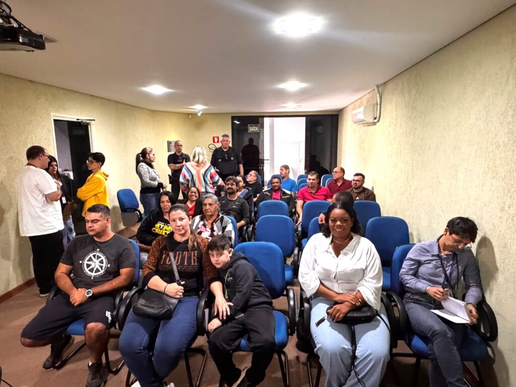 Esta é uma foto que parece ter sido tirada em uma sala de reuniões, palestras ou auditório pequeno, com várias pessoas sentadas e outras de pé. Aqui estão os detalhes que observei: O Ambiente: É uma sala com paredes bege, iluminação embutida no teto e fileiras de poltronas azuis estofadas, típicas de auditórios. Ao fundo, há uma porta de saída sinalizada. As Pessoas: Na primeira fileira (sentados): Vemos um grupo de pessoas, incluindo um homem com uma camiseta de estampa geométrica, uma mulher com uma bolsa tiracolo e um menino ao lado dela, uma mulher sorridente com uma camisa branca, e outra mulher com uma blusa azul social, que parece estar segurando ou lendo papéis. Mais ao fundo: Outras pessoas estão sentadas observando. Também há um grupo de pessoas em pé na parte esquerda e no centro da sala, incluindo alguns homens que parecem estar uniformizados, talvez parte da segurança ou organização do evento. O Clima: A cena passa a impressão de ser uma reunião comunitária, um curso, ou um evento informativo, onde as pessoas estão aguardando o início ou acompanhando uma apresentação. O ambiente é tranquilo e focado.