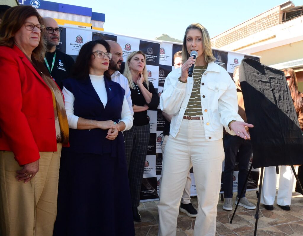 A imagem mostra um grupo de pessoas em um evento oficial do Governo do Estado de São Paulo. Ao centro, uma mulher de jaqueta branca e camiseta listrada fala ao microfone, enquanto outras pessoas — vestidas de forma formal ou semiforme — a observam atentamente. Ao fundo, há um painel com o brasão do governo e uma estrutura coberta por um tecido preto, sugerindo uma possível cerimônia de inauguração ou anúncio público.