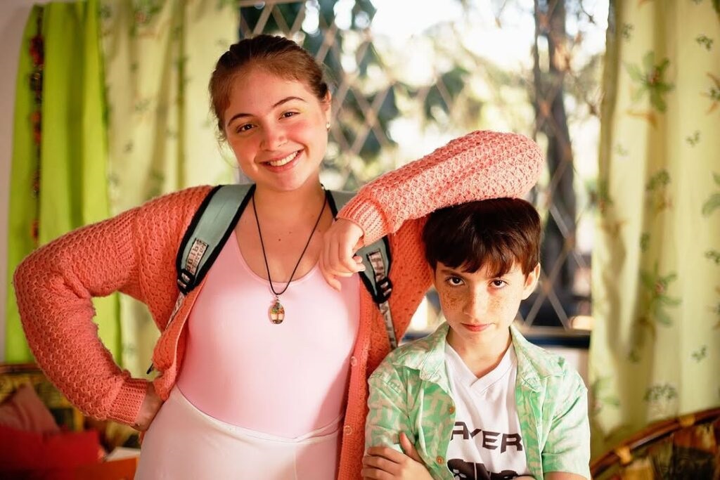 Nesta foto, temos dois jovens posando para a câmera. A garota: Está à esquerda, sorrindo abertamente. Ela usa um cardigã de tricô coral sobre um top rosa claro, uma mochila nas costas e um colar com pingente. Ela descansa o braço sobre a cabeça do menino ao lado. O menino: Está à direita, com uma expressão mais séria e um pouco de sardas no rosto. Ele veste uma camiseta branca com uma estampa escrito "PLAYER" e uma camisa aberta por cima, em tons claros de verde. Ambiente: Eles estão em um espaço iluminado, possivelmente uma sala, com uma cortina verde claro ao fundo e uma janela com grades, sugerindo um ambiente residencial tranquilo. A cena transmite uma sensação de proximidade e descontração familiar.