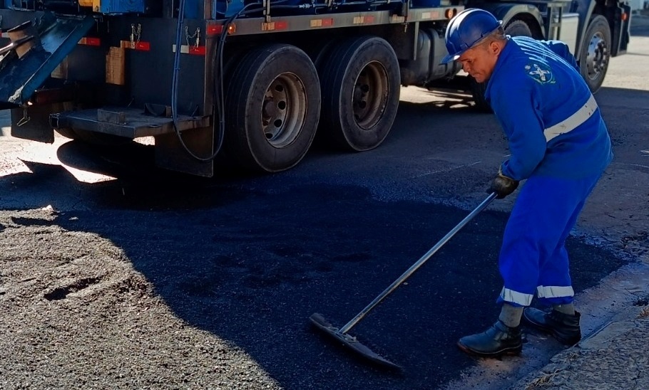 Foto de um trabalhador da manutenção viária nivelando asfalto fresco junto ao meio-fio. Detalhes observados: Homem vestido com macacão azul de segurança e capacete azul; o macacão tem faixas refletivas claras na altura do peito e das canelas. Ele usa luvas e botas de segurança e segura um rastelo/metálico longo, espalhando o asfalto. Há um caminhão ou usina de pavimentação atrás dele, com grandes pneus traseiros visíveis e parte da caçamba/mecanismo de descarregamento. O solo à frente está com asfalto escuro recém-aplicado; partes ainda parecem irregulares enquanto o trabalhador nivela. Iluminação diurna com sombras bem definidas indicando sol forte; cena provavelmente numa rua ou via urbana junto ao meio-fio. Ambiente limpo de obstruções visíveis; não há outras pessoas próximas na área mostrada.