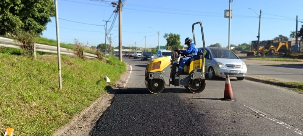 Uma cena diurna de reparo de pavimento urbano, com foco em um rolo compactador amarelo e cinza que está passando sobre um remendo de asfalto recém-aplicado à margem direita da pista. Operador: uma pessoa vestindo macacão azul e capacete azul está sentada no rolo, dirigindo-o. Trabalho: faixa estreita de asfalto novo, ainda bem escuro e granular, ocupa a borda direita da via; há um cone laranja posicionado à frente do rolo sinalizando a obra. Tráfego: ao fundo, carros seguem pela pista principal; um automóvel prata está bem próximo do rolo, ultrapassando a área de serviço. Equipamento e entorno: ao fundo à direita há mais máquinas (retroescavadeira) e um canteiro gramado à esquerda com guard-rail; postes e fios cruzam o céu limpo e azul. Iluminação e clima: sol forte, sombras definidas, clima claro — sugere manhã ou tarde ensolarada. Sensação geral: operação organizada de remendo viário em área urbana, com tráfego parcialmente desviado e sinalização simples.