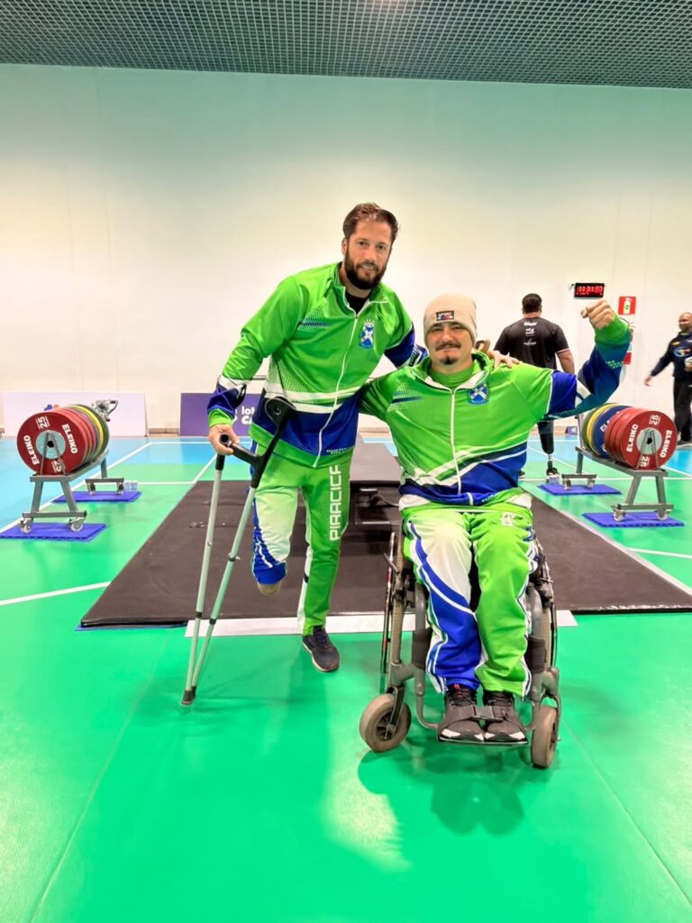 Visão geral: dois atletas posam em uma área esportiva coberta, provavelmente um ginásio para levantamento de peso adaptado. Ambos vestem agasalhos esportivos verdes com detalhes em azul e branco, formando uniforme de equipe. Posições e comportamento: À direita, um atleta sentado em cadeira de rodas sorri e ergue o braço direito em gesto de comemoração (punho fechado). Usa touca clara. À esquerda, um atleta de pé apoiado em muletas e com uma prótese na perna esquerda apoia a mão no ombro do companheiro, também sorrindo. Ele apoia uma das muletas no chão e demonstra equilíbrio apoiado na perna prótese. Ambiente e equipamentos: Ao fundo, duas plataformas de levantamento de peso com rolos de anilhas coloridas apoiadas em suportes — anilhas vermelhas com a marca “ELEIKO” são visíveis. Piso esportivo verde e uma área elevada/preparada ao centro (plataforma preta) onde provavelmente ocorrem as provas. Painel de cronômetro/placar ao fundo direito e outras pessoas em segundo plano, fora de foco. Detalhes visuais: Uniformes têm logotipo no peito e a palavra “PIRACIC” visível na perna da calça do atleta em pé. A cadeira de rodas é manual; a prótese e as muletas indicam modalidades adaptadas. Iluminação interna forte e teto com grade metálica.