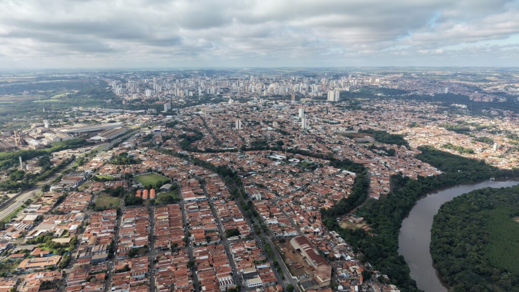 Esta é uma foto aérea impressionante de uma grande cidade, capturada em um dia com céu parcialmente nublado. Aqui estão os destaques principais: Centro Urbano: Ao fundo, vê-se uma área central densa com vários prédios altos e edifícios comerciais, destacando-se no horizonte. Área Residencial: Uma vasta extensão de bairros residenciais com telhados de cerâmica (vermelhos/laranjas) ocupa a maior parte do primeiro e segundo plano, mostrando uma urbanização bem consolidada. Natureza: À direita, um rio sinuoso corta a paisagem, ladeado por uma área de vegetação densa e preservada, o que cria um belo contraste com o cinza e o vermelho da cidade. Indústria: À esquerda, é possível observar uma área que parece industrial ou com infraestrutura de grande porte, possivelmente galpões ou complexos fabris, próximos a uma via de tráfego. É uma visão ampla e detalhada que mostra bem como a cidade se integra entre o desenvolvimento urbano e os recursos naturais.