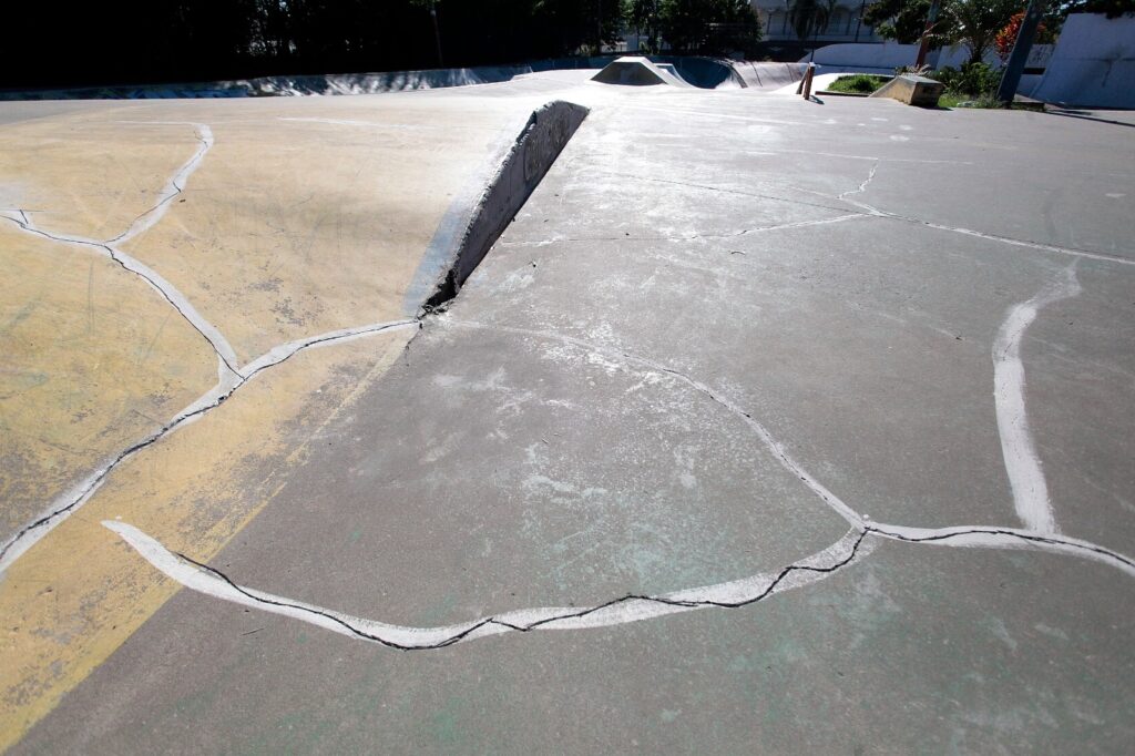 Uma pista de skate de concreto vista de perto.
No primeiro plano, o piso mostra várias fendas preenchidas com selante branco que formam linhas irregulares e curvas sobre a superfície áspera.
À esquerda há uma área com pintura amarelada desgastada; as fissuras também cortam essa região.
Ao centro, em direção ao fundo, aparece um pequeno obstáculo de concreto (um bisel/ombro) com a junção onde o selante começa.
Mais ao fundo vê-se a continuação da pista com rampas e transições suaves, e um relevo que parece uma pequena rampa triangular.
Na borda direita e ao fundo há vegetação e algumas sombras de árvores, além de um muro branco ao longe.
A cena está iluminada por luz forte do sol, criando brilho sobre o concreto e contrastes nítidos entre áreas claras e sombreadas.
A imagem transmite um espaço usado e um pouco gasto, típico de um skate park ao ar livre.